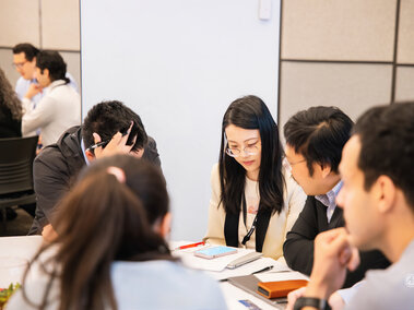Group of people seated at a table, focused on paperwork and discussion in a bright room.
