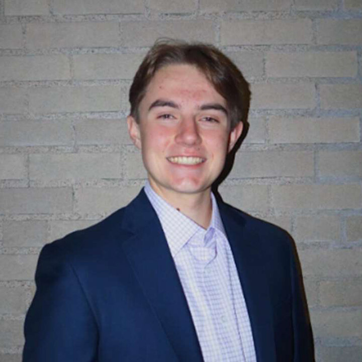 Person in a navy blazer and checkered shirt standing against a beige brick wall.