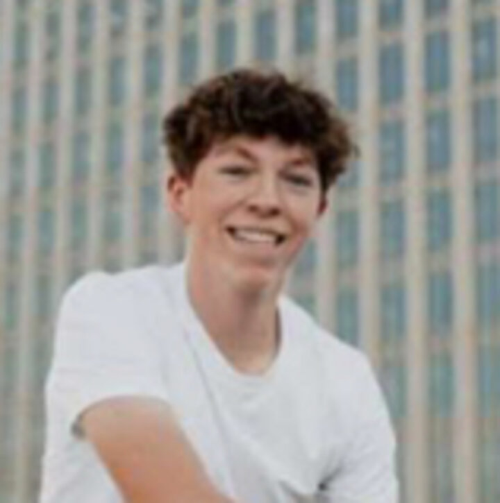 Young person in a white shirt, seated outdoors, with a tall building in the background.