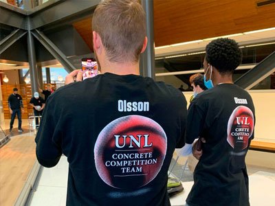 Two students stand with their backs to the camera with shirts that say "UNL Concrete Competition Team"