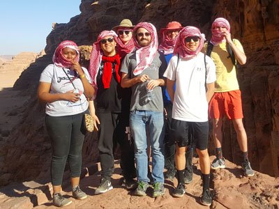 Seven people stand on a canyon wearing red scarves to protect their heads from the sun and sand