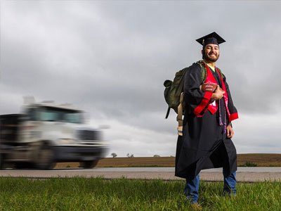 Man stands outside in a black graduation gown holding a backpack