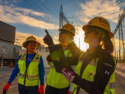 Three women wear hard hats and construction vests on a work site.