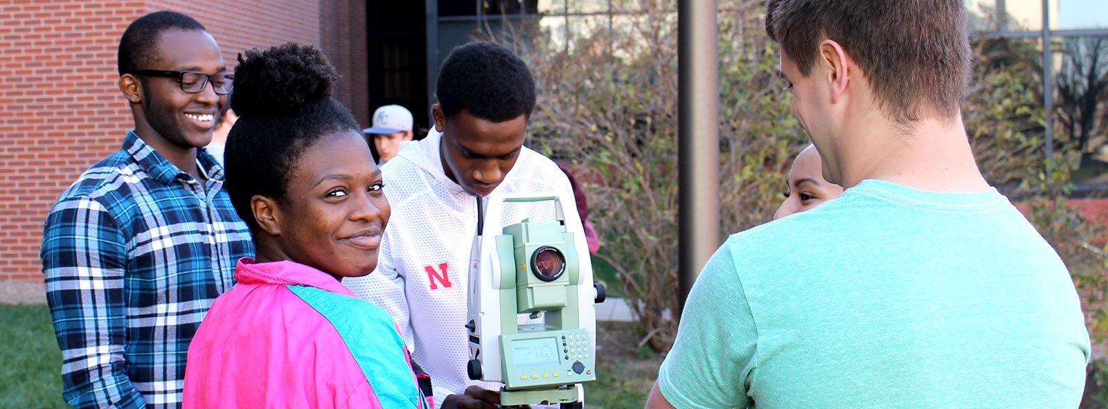 Four engineering students use a scope to survey outside the engineering complex. One student is facing the camera and smiling and another is looking into the scope.